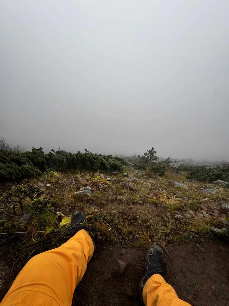 Yuzhe's view past yellow hiking trousers and boots toward a rocky, mist-covered hillside.