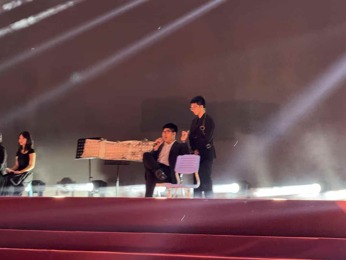 Yuzhe seated on stage in a dark suit, holding a microphone, with other performers nearby.